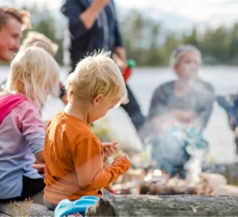 En grupp människor, inklusive ett barn, sitter runt en lägereld ute i naturen vid Orsa Grönklitt. De njuter av en avkopplande stund och lågan sprakar i förgrunden.