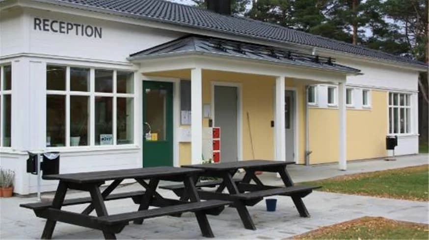 Bilden visar en campingplats med en grön gräsmatta och flera utomhusbänkar i skuggan. I bakgrunden syns en byggnad med dörrar och träpaneler, som kan vara en del av campinganläggningen.