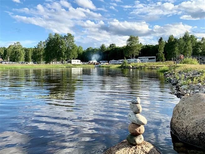 En somrig campingplats vid sjön med frodig grönska och ett lugnt vattendrag som strömmar genom området, omgiven av träd och öppna ytor för rekreation och avkoppling.