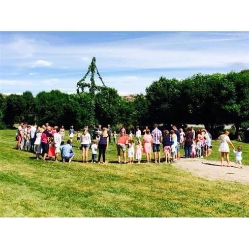 En grupp människor, inklusive barn, njuter av en solig dag på Wiggersviks camping, omgivna av frodig grönska. Några står i klungor och pratar, medan andra leker eller kopplar av. I bakgrunden syns tält och campingutrustning. Stämningen är livlig och avslappnad, med många engagerade i olika fritidsaktiviteter.
