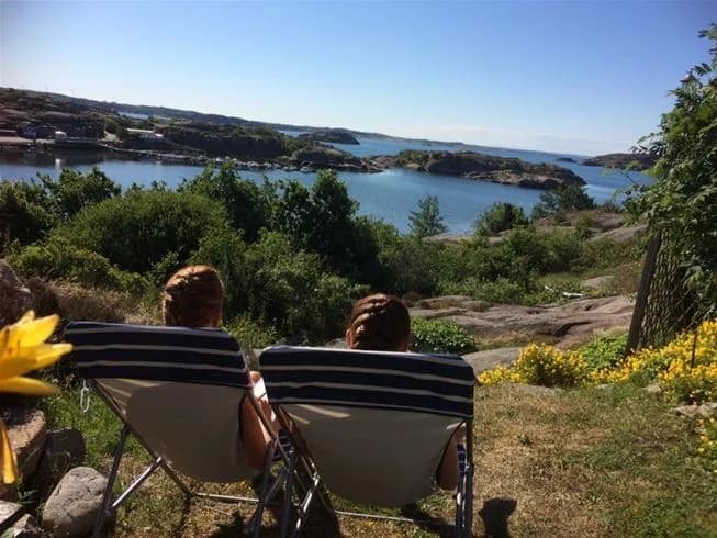 En naturskön vy vid Wiggersviks camping med utomhusmöbler för picknick, omgivande kustlandskap och en vik i bakgrunden.