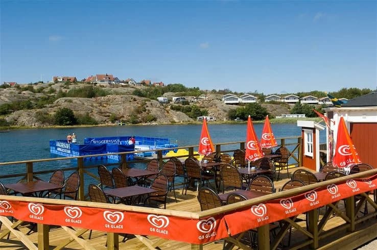 En bild av Wiggersviks camping med båtar vid strandkanten, utomhusmöbler och en restaurang i bakgrunden, vilket ger en inbjudande känsla av semester i en kustnära småstad.