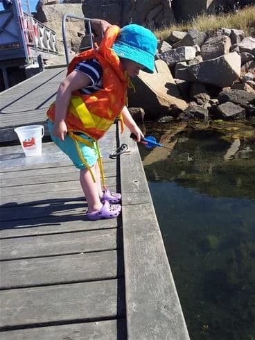 Ett barn i sneakers står på en brygga och fiskar, med campingen Wiggersviks som bakgrund, vilket ger en känsla av somrig rekreation och semester.