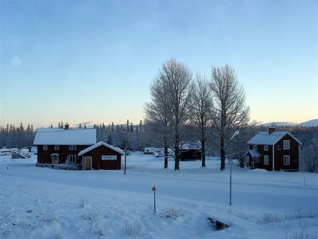 En vinterbild med snötäckta trädgrenar framför Vålågårdens värdshus & camping, ett mysigt hus med snö på taket beläget i en sluttning.
