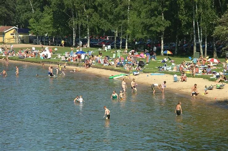 En solig dag vid Vadstena Camping med en sjö i bakgrunden där människor njuter av fritidsaktiviteter vid stranden.