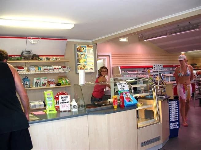 En kund står vid en hylla fylld med livsmedel på Vadstena Campings butik, där en expedit hjälper till. Inredningen påminner om en snabbköpsmiljö.