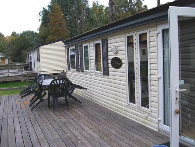 En husbil står parkerad vid sidan av en trädäckad uteplats på Vadstena camping. I bakgrunden syns frodig grönska och en klarblå himmel.