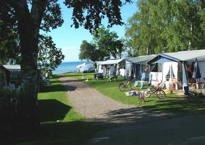 Bild av Vadstena camping med utsikt över kusten. Stranden är synlig med skugga från träd. Utemöbler finns för rekreation vid havet, och en vik breder ut sig i bakgrunden.