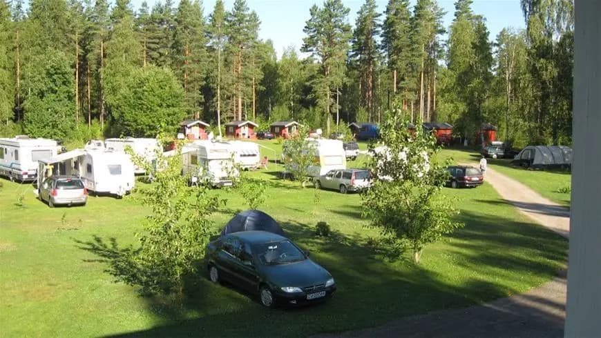 En grön campingplats omgiven av träd och växter vid Sörälgens Camping, med gräsmatta och parkeringsytor för husbilar och fritidsfordon, i en naturskön miljö idealisk för rekreation.