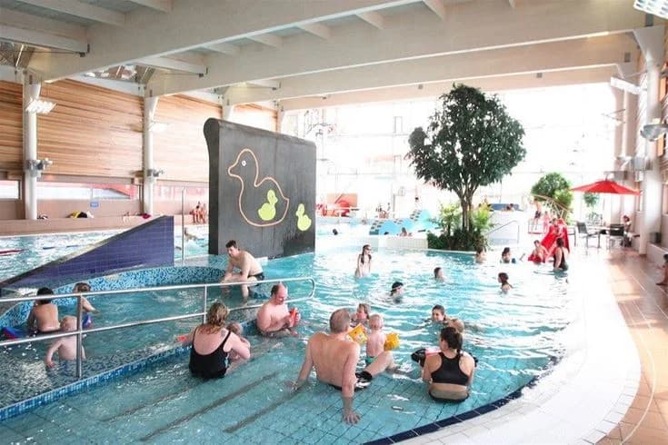 En livlig campingplats med gäster som njuter av bad och rekreation vid en pool. Några besökare bär badkläder och deltar i olika fritidsaktiviteter, vilket bidrar till en avslappnad semestermiljö. I bakgrunden syns fler delar av campingen och omgivande natur.