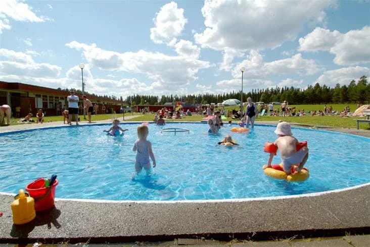 En solig dag vid Strömsunds camping, där besökare njuter av en uppfriskande simtur i utomhuspoolen, omgivna av gröna träd och en avslappnad semesteratmosfär.