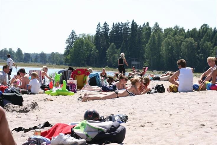 En solig sommardag på Strandstuvikens havsbad & camping, där människor njuter av stranden, badar och solar på sanden iförda badkläder.
