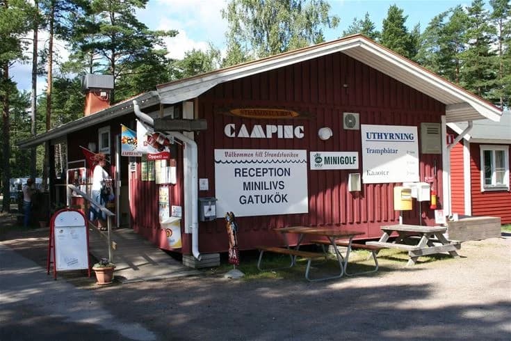 En solig dag vid Strandstuvikens havsbad & camping, med en trästuga omgiven av träd som ger skugga. En avkopplande rastplats i naturlig miljö.