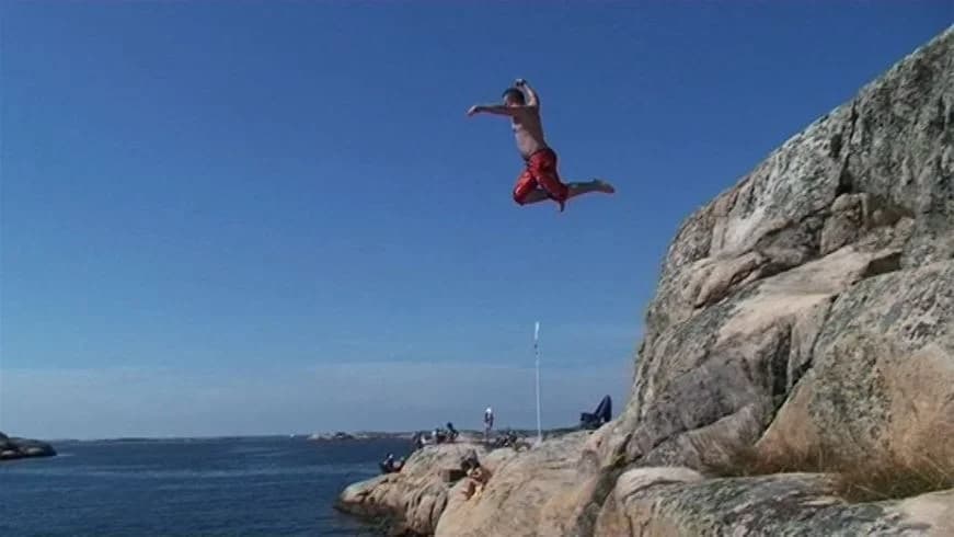 Person som hoppar från klippa ner i vattnet vid kustnära terräng nära Sotenäs Camping, med havet i bakgrunden.