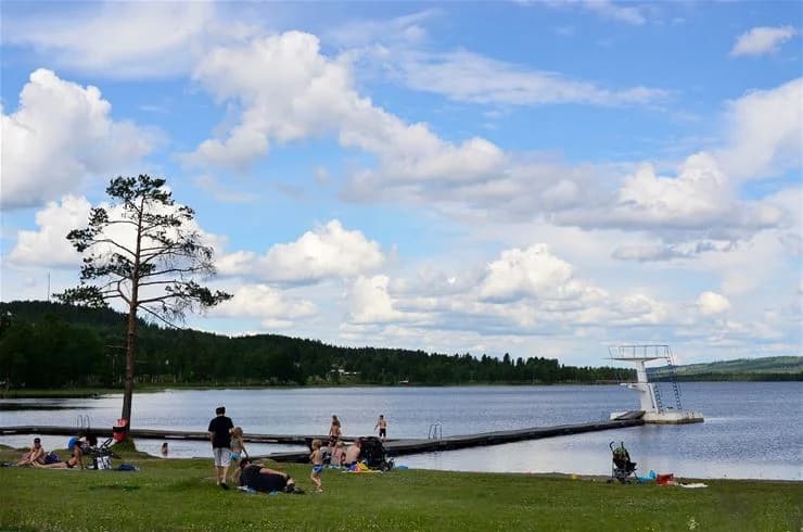 Vy över Saiva Camping & Stugby med en sjö i bakgrunden under en klar himmel. Människor njuter av fritidsaktiviteter och semester vid vattnet, omgivet av natur.