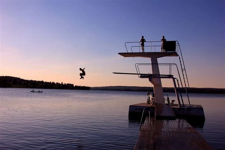 En person hoppar från en brygga ner i vattnet vid solnedgången på Saiva Camping & Stugby. Atmosfären är lugn, och himlen är färgad i varma toner av kvällsskymningen.