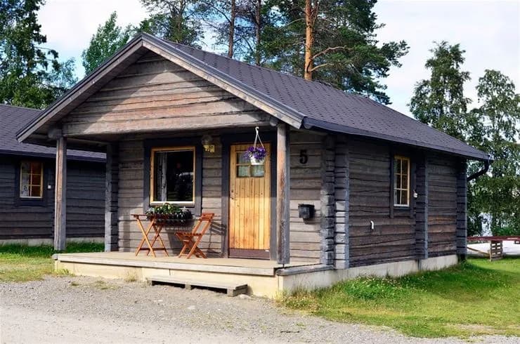 En lägerplats med flera timmerstugor belägna i en lantlig miljö, omgivna av grönska. Stugorna har trätak och dörrar, vilket skapar en hemtrevlig och rustik atmosfär.