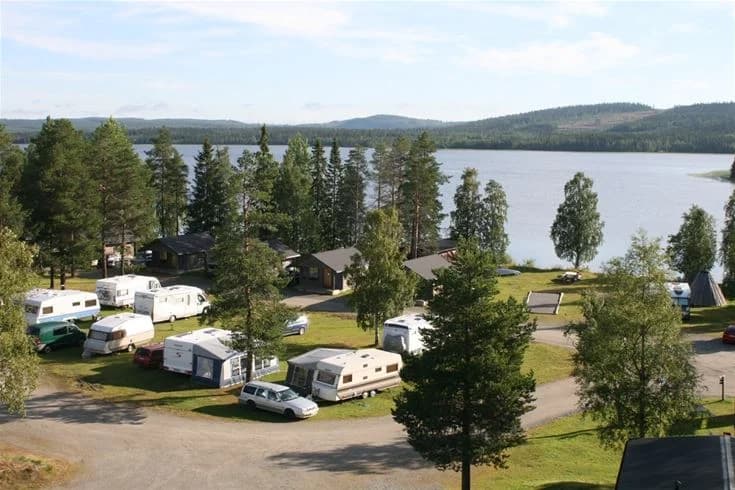 En lugn sjö omgiven av tät barrskog med granar och lärkträd, med parkerade fordon vid Saiva Camping & Stugby.