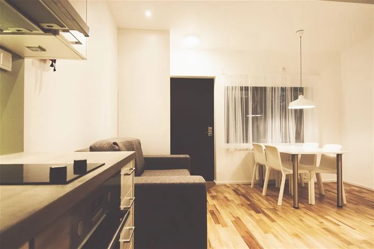 Ett interiört rum med trägolv och vita väggar. Rummet är möblerat med enkla trä- och tygmöbler. Taklampor hänger från det vita taket, och en mjuk belysning skapar en inbjudande atmosfär.