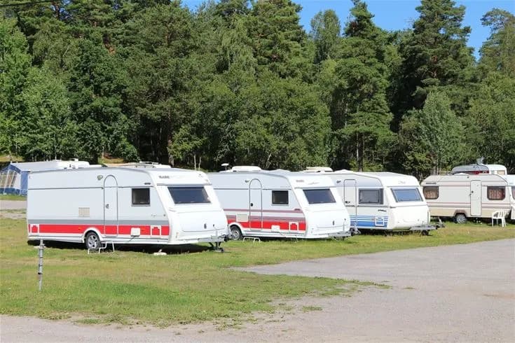 En husvagn står parkerad på en stor grusplan omgiven av träd, med flera andra husvagnar och bilar synliga i bakgrunden.