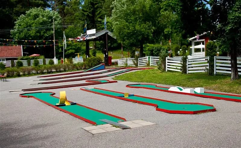 En lekplats med konstgräs som inkluderar utrustning för utomhuslek samt en minigolfbana, beläget i en parkmiljö vid Rösjöbaden Camping & Stugby.