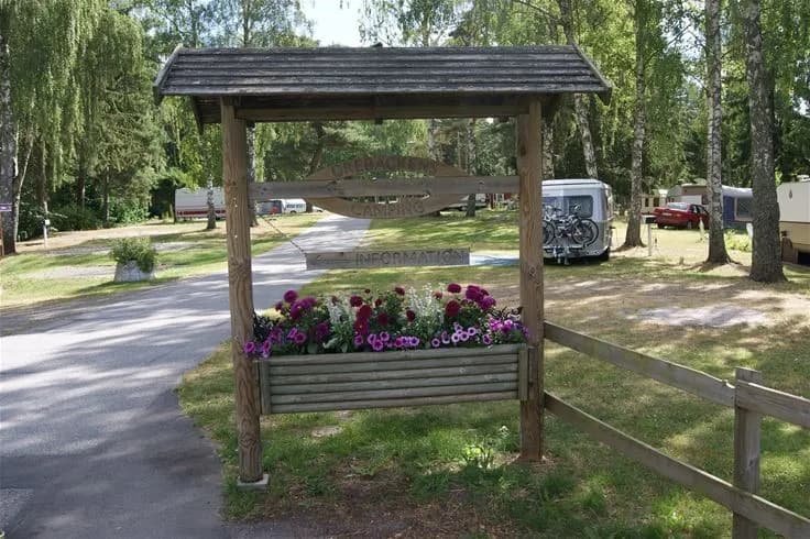 En lummig trädgård med skuggiga områden på Orebackens camping, omgiven av välskötta planteringar och grönytor. I mitten syns en trädgårdspaviljong med sittplatser, idealisk för avkoppling.