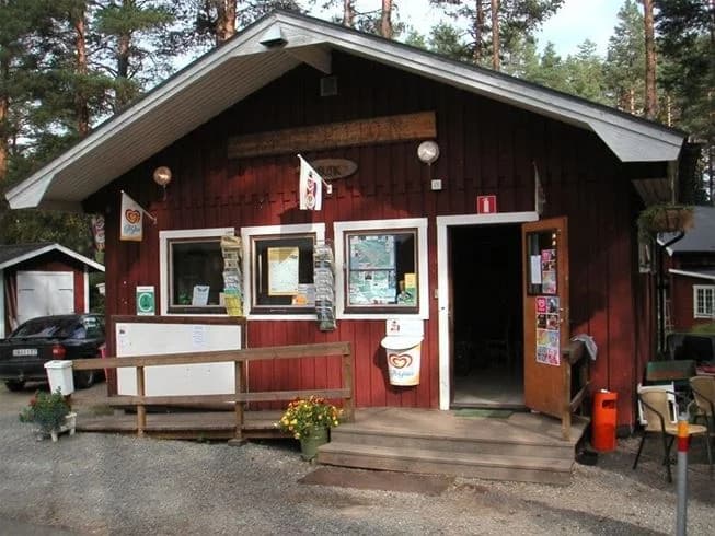 En trästuga med dörr och skugga omgiven av en välskött gård, trädgårdsbyggnader och en bänk i rastområdet.