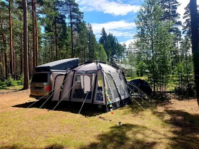 En campingplats med flera husbilar och minibussar parkerade på grusunderlag. Omgivningarna är grönskande med träd i bakgrunden, vilket ger en känsla av fridfullhet och närhet till naturen.