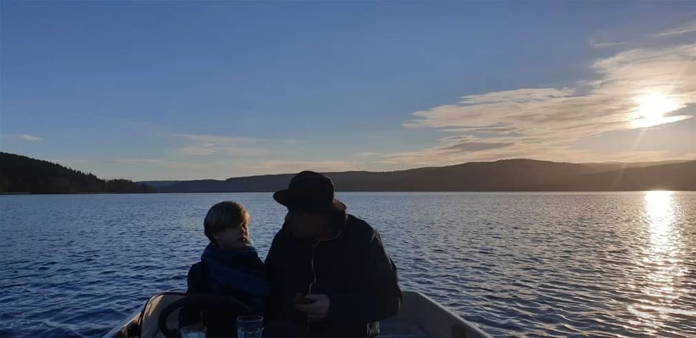 En stillsam sjö i skymningen med en båt som flyter på vattnet, himlen är delvis täckt av moln vid horisonten. Naturcamping Lagom.