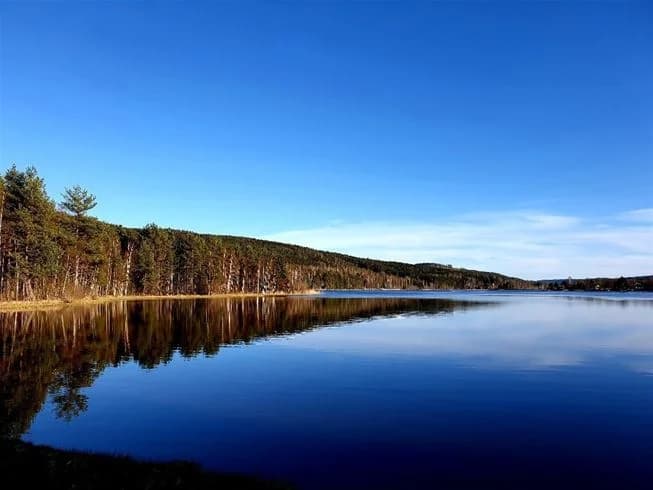 En blå himmel sträcker sig över en spegelblank vattenyta vid Naturcamping Lagom, med naturlandskap synligt vid horisonten.