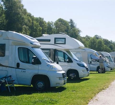 En bild som visar en husvagn parkerad på Marstrands Familjecamping med flera bilar runtomkring. Flera husvagnar kan ses i bakgrunden på den öppna campingplatsen.