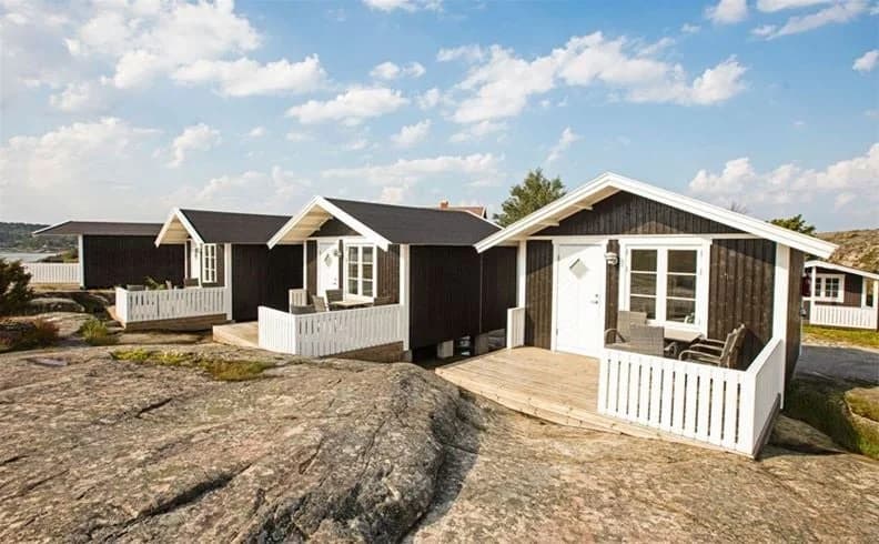 En bild av en pittoresk stuga med sluttande tak belägen vid Lökholmens camping, omgiven av grönska och med närhet till andra små byggnader i en lugn bymiljö.