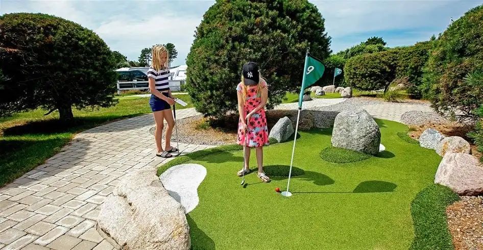 En person spelar minigolf på Lökholmens camping. Gräsplan omgiven av träd och buskar i bakgrunden. Flera färgglada golfbollar och klubbor syns på banan. Avkopplande utomhusmiljö.