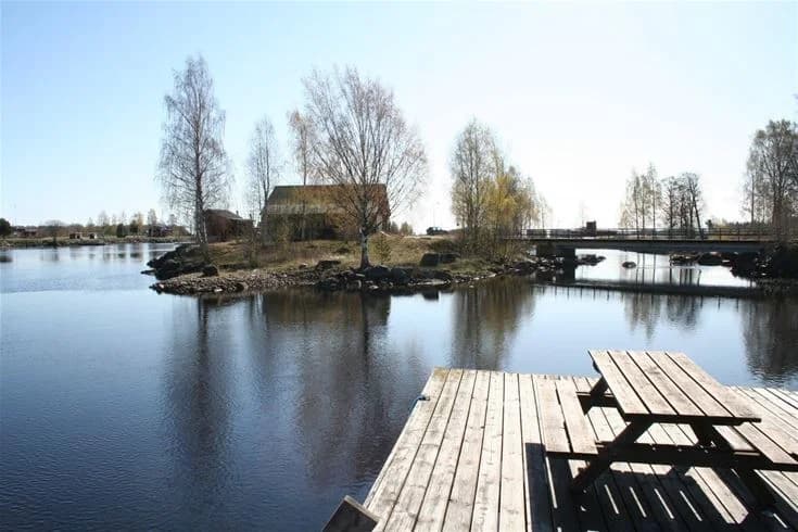 En stilla sjö vid Ljusnefors camping omgiven av lummiga träd och naturlandskap, med en smal vattenkanal längs strandkanten.