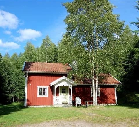 En mysig stuga omgiven av grönska på Jälluntofta camping, med träd i bakgrunden och en välskött gräsmatta i förgrunden.