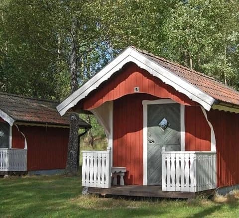 En liten trästuga med mörkt tak, omgiven av grönska och flera små trädgårdsbyggnader, på Jälluntofta camping.