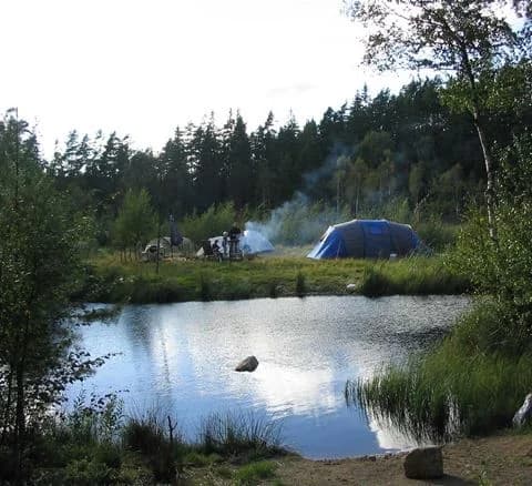 En tältplats omgivet av träd och grönska vid en liten damm i Hätteboda vildmarkscamping. En tarpaulin är uppsatt över tältområdet.