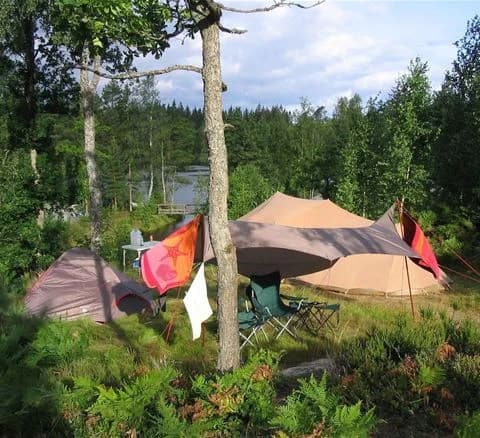 En campingplats på Hätteboda Vildmarkscamping med ett uppställt tält och presenning för skugga. Campingen är utrustad med utomhusmöbler och friluftsutrustning, vilket skapar en inbjudande atmosfär för friluftsliv och semester.