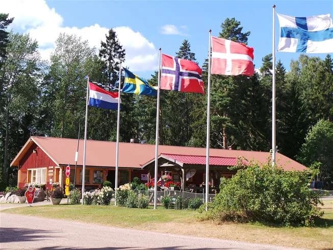 En campingplats med träd som ger skugga, en flaggstång med en amerikansk flagga, och människor som njuter av fritidsaktiviteter i området.