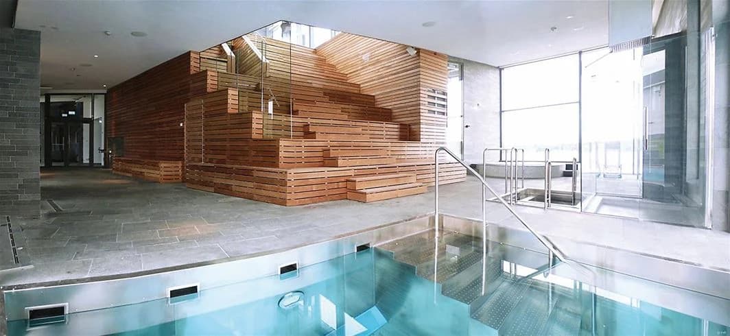 En modern inomhusmiljö med en pool omgiven av glasväggar och trägolv. Ljuset strömmar in, vilket skapar en ljus och avkopplande atmosfär.