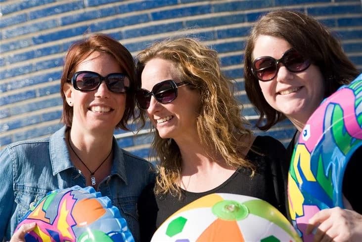 En grupp glada människor med solglasögon njuter av solen på Bovikens havsbad. Bakgrunden visar en sandstrand och havet.