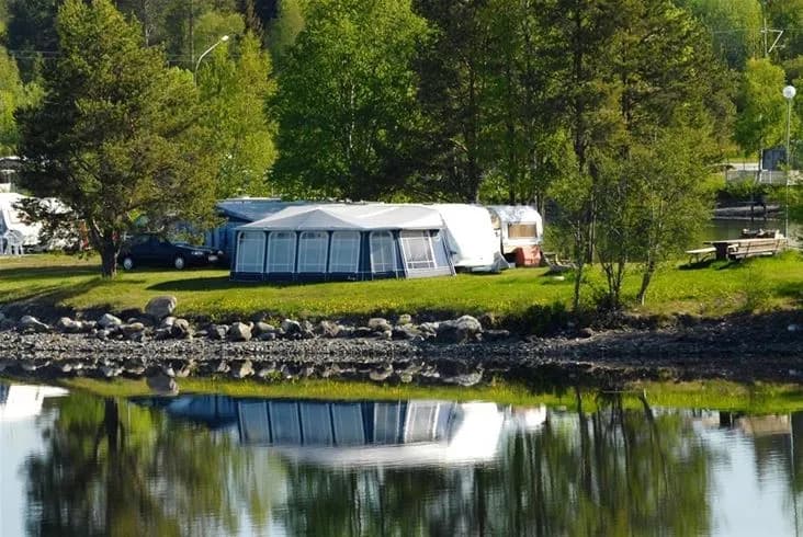 Alt-text: Lugn campingplats vid vattnet i Krokomvikens camping med tält uppställda vid en sjö omgiven av grönska och flodlandskap.
