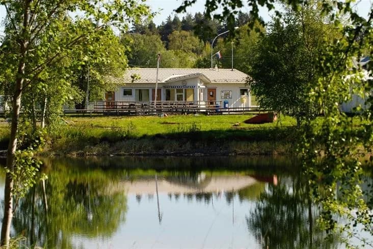 En vy över Krokomvikens camping med en stilla vattenspegel omgiven av träd och växtlighet, där vattnet vackert reflekterar den omkringliggande naturen.