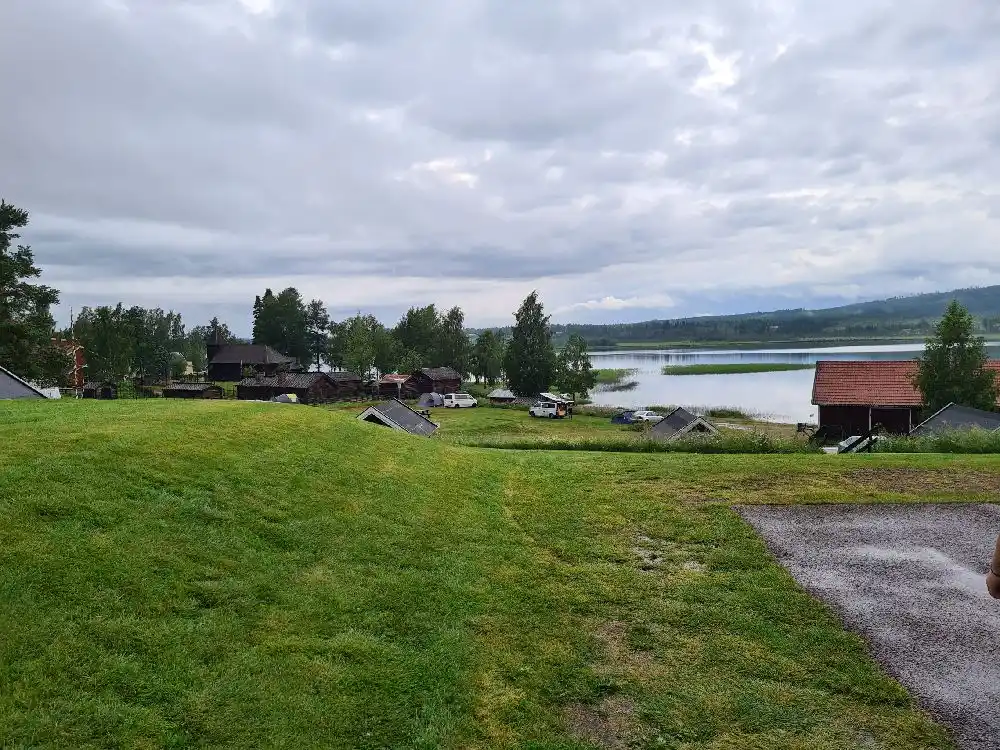 En vidsträckt campingen vid Särna, omgiven av grön gräsmark under en himmel med vita, fluffiga cumulusmoln. I bakgrunden syns den lantliga, öppna naturen med åkrar och betesmarker, vilket skapar en lugn och naturnära atmosfär.