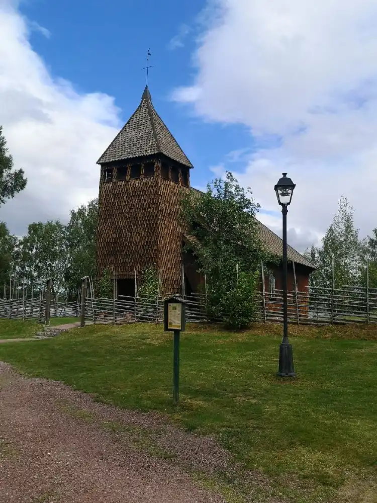 En campingplats omgiven av ett trästaket, med en kyrkspira synlig i bakgrunden under en klar himmel.