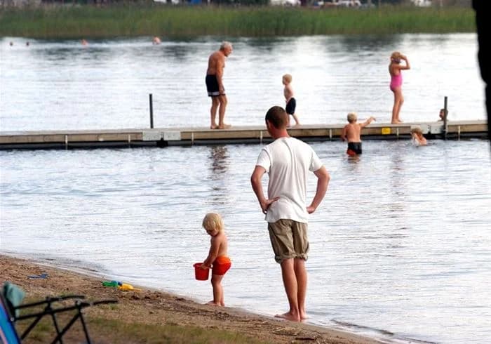 En grupp människor njuter av sommaraktiviteter vid sjön på Karlsborgs camping, några vandrar barfota i strandkanten och andra kopplar av i solen med lek och rekreation.
