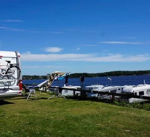 En campingplats med utsikt över en marina där flera båtar ligger förtöjda. I förgrunden syns parkerade husbilar och husvagnar nära vattnet.