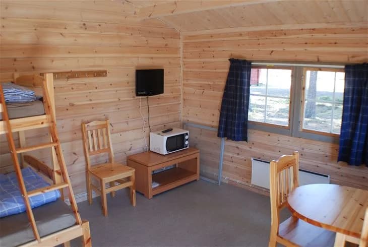 En trästuga vid Storuman camping, omgiven av skog och andra trädbyggnader, med träplankor och vedstaplar synliga i förgrunden.