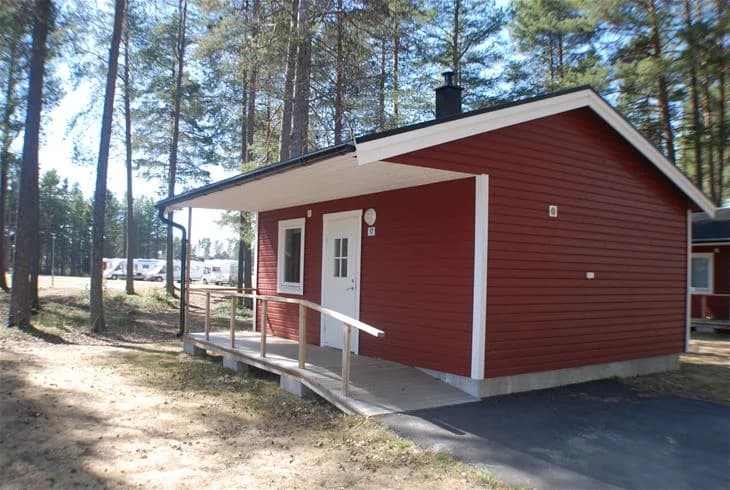 En timmerstuga med träfasad och en dörr, omgiven av en grönskande trädgård på Storumans camping.