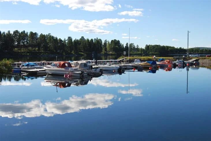 En småbåtshamn med flera motorbåtar ligger förtöjda vid bryggorna på Storumans camping. Vattnet är lugnt och speglar båtarna och det omgivande landskapet.